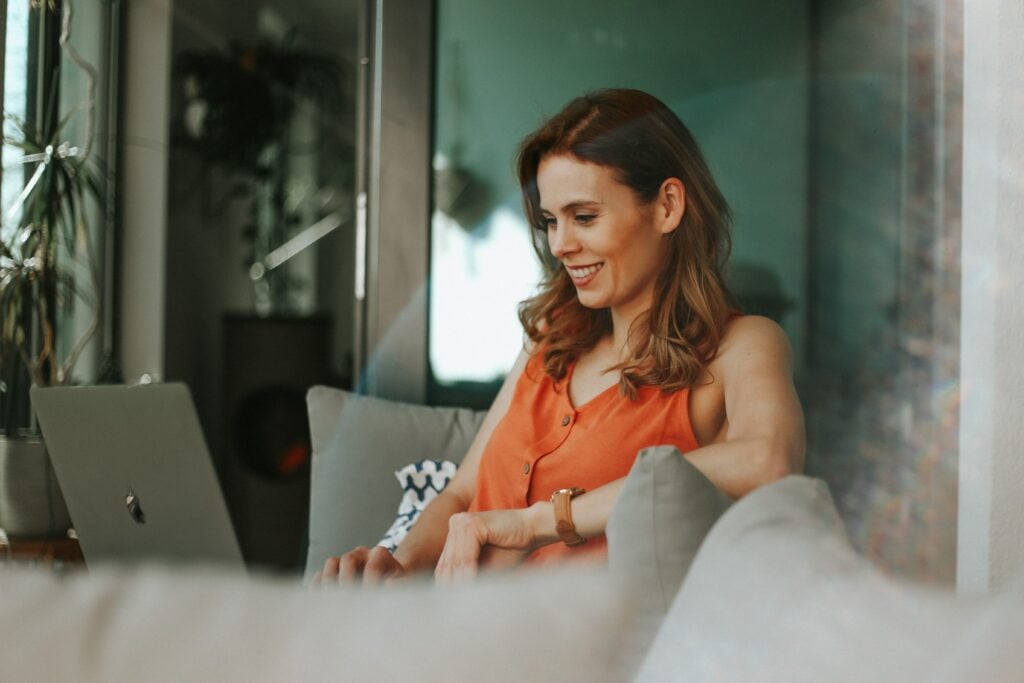 Woman in orange top grins at laptop.