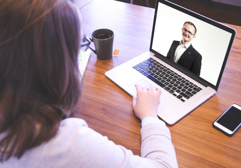Image of a woman with a cup of tea, looking at a screen with a man wearing glasses and suit smiling on it
