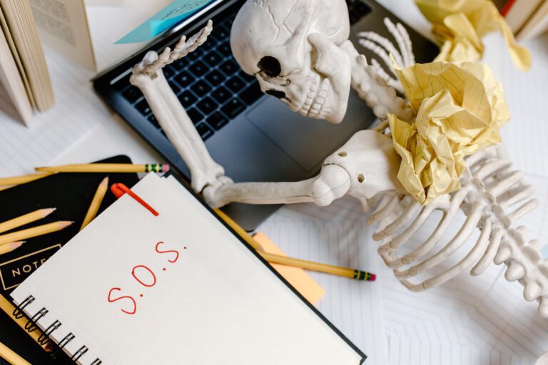 Bekämpfung von remote work burnout Bild eines Skeltons, der an einem Computer sitzt und SOS auf ein Notizbuch neben sich schreibt
